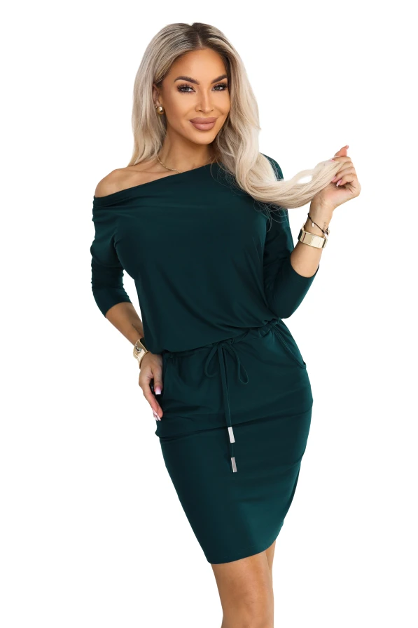 Model purtând o rochie sport de culoarea verde sticlă, cu decolteu ce dezvăluie un umăr, mâneci 3/4, buzunare laterale și legătură la talie. Croi cu partea de sus lejeră și partea de jos mulată, chiar deasupra genunchilor.