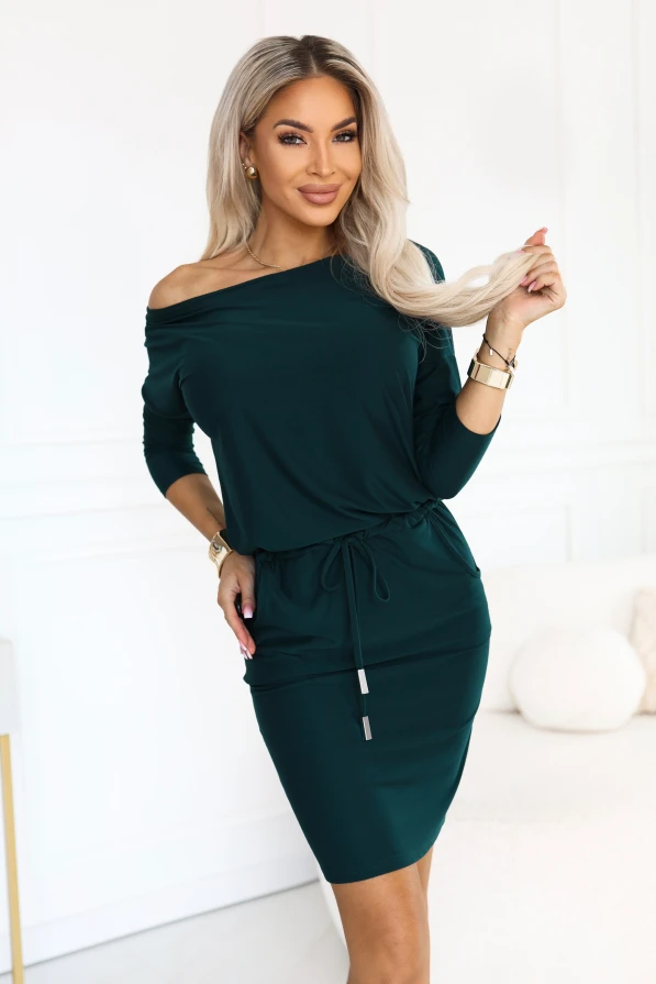 Model purtând o rochie sport de culoarea verde sticlă, cu decolteu ce dezvăluie un umăr, mâneci 3/4, buzunare laterale și legătură la talie. Croi cu partea de sus lejeră și partea de jos mulată, chiar deasupra genunchilor.