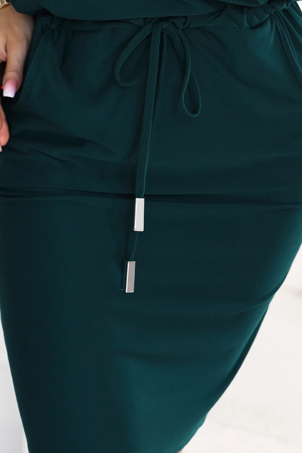 Model purtând o rochie sport de culoarea verde sticlă, cu decolteu ce dezvăluie un umăr, mâneci 3/4, buzunare laterale și legătură la talie. Croi cu partea de sus lejeră și partea de jos mulată, chiar deasupra genunchilor.