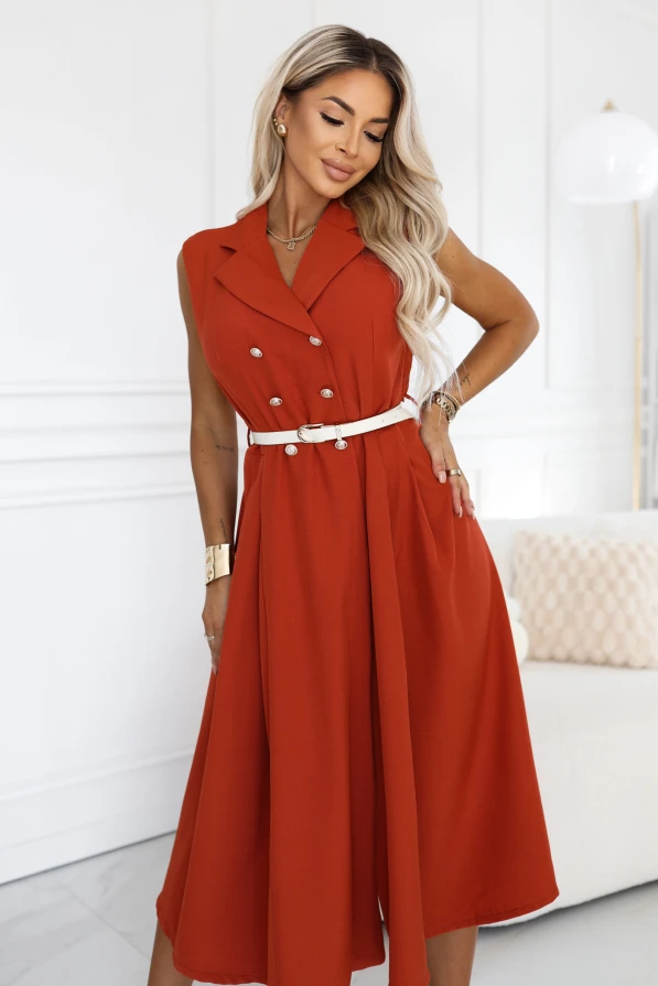 Modelka w ceglastej sukience marynarkowej midi marki Numoco Basic. Fason bez rękawów ma szerokie klapy i dwa rzędy ozdobnych guzików, a talię podkreśla wąski, jasny pasek.