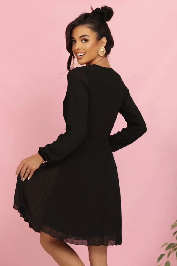 Modelka prezentuje elegancką czarną szyfonową sukienkę marki Numoco z długim rękawem, kopertowym dekoltem i plisowanym dołem – idealną na wieczór, wesele lub specjalne okazje. Produkt polski, sfotografowany na różowym tle.