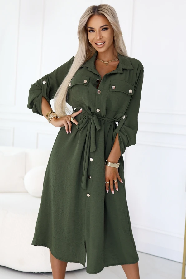 Stylowa sukienka koszulowa w kolorze khaki, z wiązaniem w pasie, guzikiem zapinana na całej długości, długi rękaw z podpięciem, elegancki fason marki Numoco Basic, europejska produkcja, prezentacja na modelce w nowoczesnym wnętrzu.