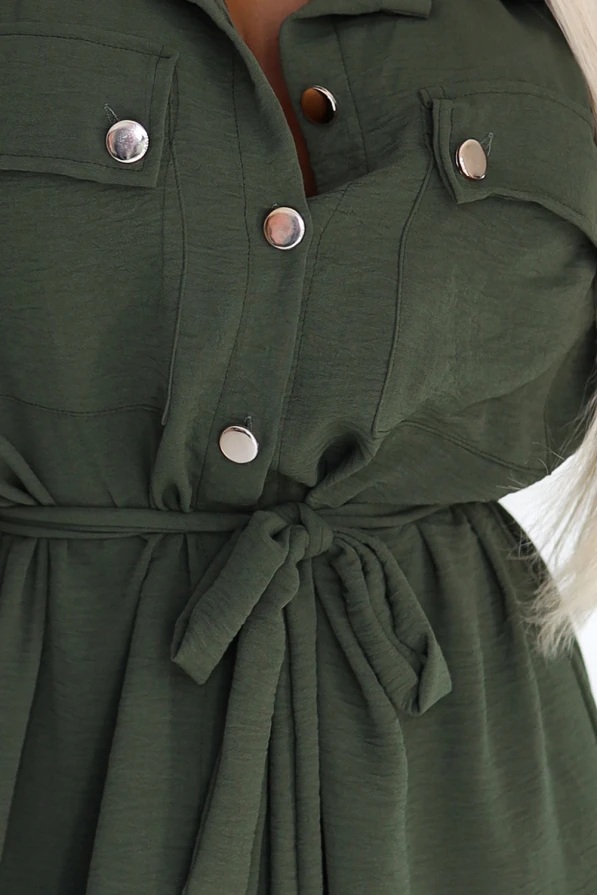 Stylowa sukienka koszulowa w kolorze khaki, z wiązaniem w pasie, guzikiem zapinana na całej długości, długi rękaw z podpięciem, elegancki fason marki Numoco Basic, europejska produkcja, prezentacja na modelce w nowoczesnym wnętrzu.